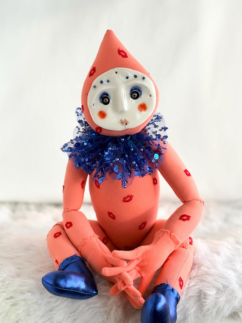 Puede incluir: Una mu&ntilde;eca caprichosa con un cuerpo color melocot&oacute;n adornado con huellas de labios rojos. Presenta una cabeza en forma de l&aacute;grima, una cara blanca en forma de m&aacute;scara con ojos azules y un cuello con lentejuelas azules. La mu&ntilde;eca lleva zapatos met&aacute;licos azules.