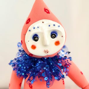 Puede incluir: Una mu&ntilde;eca hecha a mano con un dise&ntilde;o caprichoso. La mu&ntilde;eca presenta un cuerpo rosa y un sombrero en forma de cono adornado con huellas de labios rojos. Tiene una cara blanca con ojos azules, mejillas rosadas y un cuello con lentejuelas azules. El dise&ntilde;o de la mu&ntilde;eca recuerda a un buf&oacute;n o payaso.