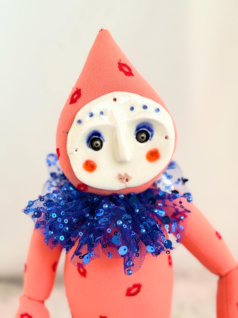 Puede incluir: Una mu&ntilde;eca hecha a mano con un dise&ntilde;o caprichoso. La mu&ntilde;eca presenta un cuerpo rosa y un sombrero en forma de cono adornado con huellas de labios rojos. Tiene una cara blanca con ojos azules, mejillas rosadas y un cuello con lentejuelas azules. El dise&ntilde;o de la mu&ntilde;eca recuerda a un buf&oacute;n o payaso.