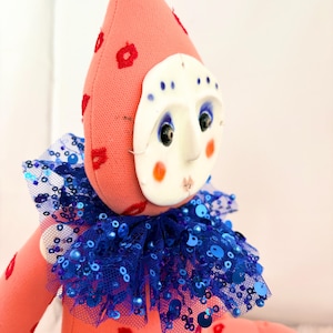 Puede incluir: Mu&ntilde;eca hecha a mano con cuerpo rosa y gorro puntiagudo adornado con motivos florales rojos. Presenta una m&aacute;scara blanca con rasgos faciales pintados y un cuello de lentejuelas azul. El dise&ntilde;o de la mu&ntilde;eca es caprichoso y art&iacute;stico.