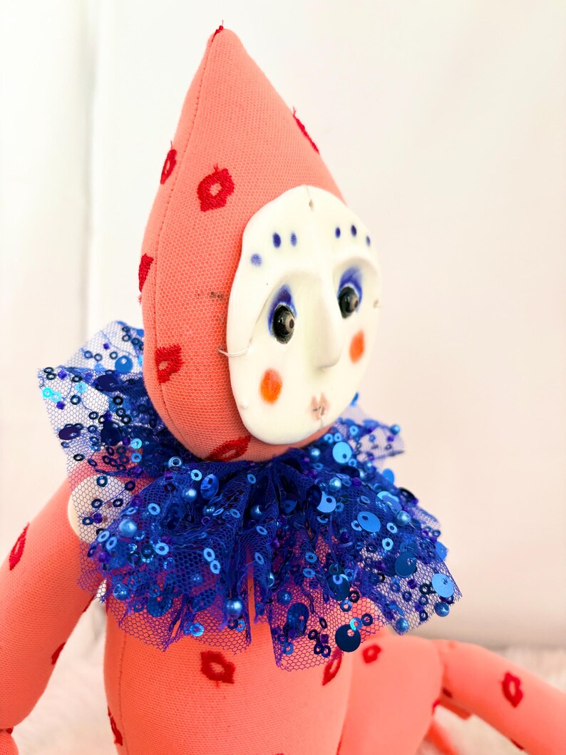 Puede incluir: Mu&ntilde;eca hecha a mano con cuerpo rosa y gorro puntiagudo adornado con motivos florales rojos. Presenta una m&aacute;scara blanca con rasgos faciales pintados y un cuello de lentejuelas azul. El dise&ntilde;o de la mu&ntilde;eca es caprichoso y art&iacute;stico.