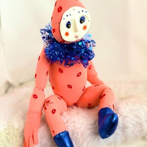 Puede incluir: Una mu&ntilde;eca artesanal con cuerpo color melocot&oacute;n y un gorro puntiagudo adornado con huellas de labios rojos. Presenta una m&aacute;scara blanca con ojos pintados y mejillas rosadas, un cuello de lentejuelas azul y botas azules. La mu&ntilde;eca est&aacute; sentada.