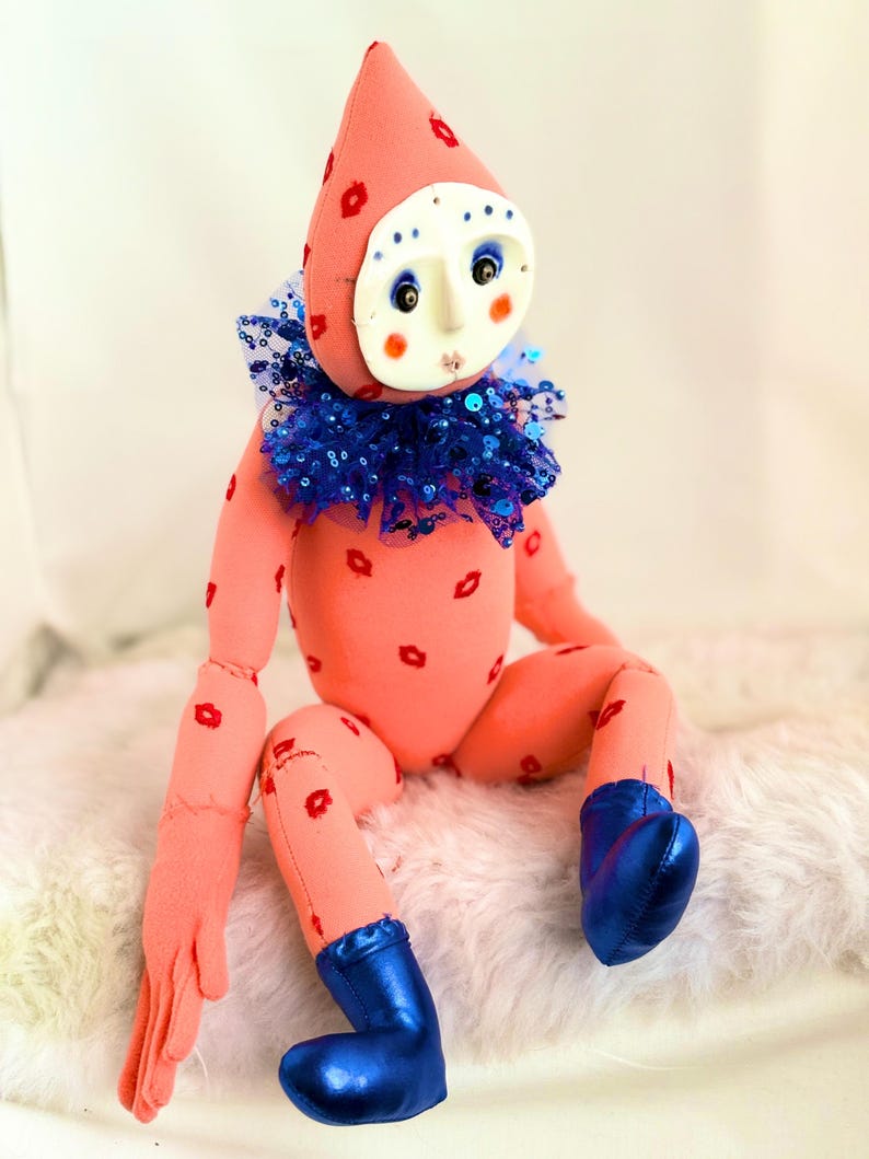 Puede incluir: Una mu&ntilde;eca artesanal con cuerpo color melocot&oacute;n y un gorro puntiagudo adornado con huellas de labios rojos. Presenta una m&aacute;scara blanca con ojos pintados y mejillas rosadas, un cuello de lentejuelas azul y botas azules. La mu&ntilde;eca est&aacute; sentada.