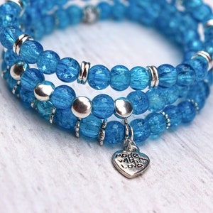 Puede incluir: Un brazalete de triple vuelta hecho con cuentas de vidrio azul y detalles plateados. El brazalete tiene un dije en forma de corazón con la inscripción "Made with Love".