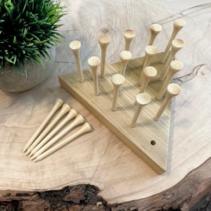 May include: A wooden triangular game board with 15 wooden golf tees standing upright in a triangular pattern. Five additional golf tees are lying on the surface next to the board.