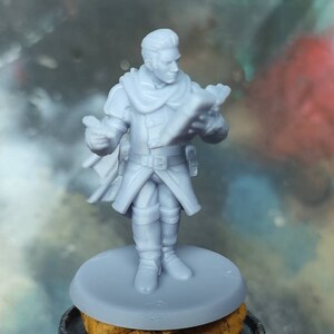 May include: A gray plastic miniature figurine of a person wearing a long coat and holding a book. The figurine is standing on a round base.