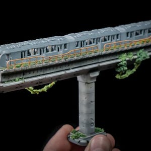 May include: A miniature gray model of a subway train on an elevated track. The model features a detailed design with windows, a yellow railing, and green foliage. The train is supported by a concrete pillar.