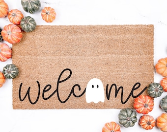 Puede incluir: Una alfombra de coco marrón con la palabra "welcome" escrita en letras negras con un fantasma blanco en el medio de la palabra. La alfombra está rodeada de pequeñas calabazas naranjas y verdes.