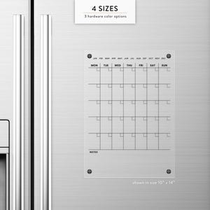 May include: A clear acrylic calendar with a grid layout, mounted on a stainless steel refrigerator. The calendar displays days of the week and months. Text at the top reads "4 SIZES" and "3 hardware color options". The size shown is 10" x 14".