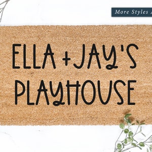 May include: A rectangular doormat made of natural coir fiber. The mat features the text "ELLA + JAY'S PLAYHOUSE" in bold, black letters. Greenery accents the corners. The words "More Styles Available" are in a blue rectangle.