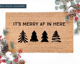 May include: A brown coir doormat with the text "It's Merry AF in Here" in black lettering. Four stylized black Christmas trees are arranged below the text.