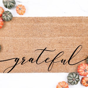 May include: A brown coir doormat with the word "grateful" written in black cursive script. The doormat is surrounded by small orange and green gourds.
