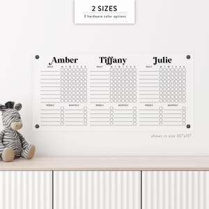 May include: A clear acrylic chore chart with three columns labeled Amber, Tiffany, and Julie. Each column has daily, weekly, and monthly sections for tracking tasks. The chart is shown in a 30" x 15" size and has text that says "2 SIZES" and "3 hardware color options."