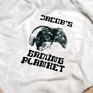 May include: White fleece blanket with black text that says "Jacob's Gaming Blanket" and a black and white illustration of a video game controller and headphones.