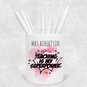 May include: White ceramic pen pot with the name "MRS ROBERTSON" and the phrase "TEACHING IS MY SUPERPOWER" in a comic book style. The design includes a pink watercolour splash and yellow stars. Several white pens are inside.