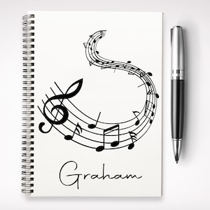 May include: A notebook with a spiral binding, featuring a musical staff design and the name "Graham". A black and silver pen is positioned to the right. The musical notes are black on a white background.