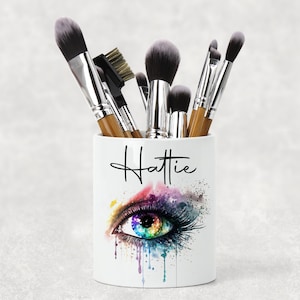 May include: White ceramic makeup brush holder with a colourful eye design and the name "Hattie" written in black script.