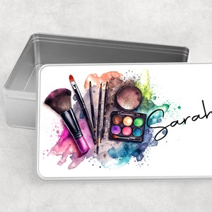 May include: A white metal tin featuring a colourful watercolour design of makeup brushes and an eyeshadow palette. The tin is inscribed with the name "Sarah" in black cursive.