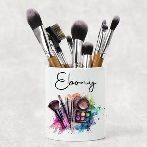 May include: White ceramic makeup brush holder with the name "Ebony" written in black cursive. The holder is decorated with a watercolour design of makeup brushes and a palette of eyeshadows.