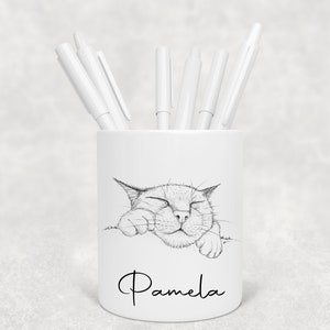 May include: White ceramic pen pot featuring a black and white cat illustration. The cat has closed eyes and paws resting on the pot's edge. The name "Pamela" is written in cursive below the cat. Several white pens are placed inside the pot.