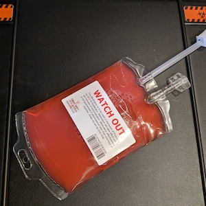 Könnte beinhalten: Ein klarer Plastikbeutel gefüllt mit roter Flüssigkeit, mit einem weißen Etikett mit der Aufschrift "WATCH OUT". Der Beutel hat einen Schlauch und einen Anschluss. Der Hintergrund ist eine schwarze Oberfläche mit orangefarbenen Gefahrenaufklebern.