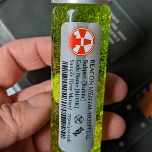May include: A small glass vial filled with a green liquid. The vial has a white label with black text that reads "BEACON MENTAL HOSPITAL Subject-[Rubin] Code Name-[RUVIK] Sample-[Grey Matter] 77501/324-221b". There is a red biohazard symbol on the label.