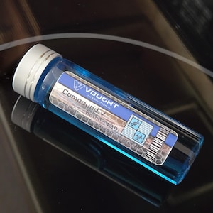 Può includere: Una bottiglia di vetro blu con un'etichetta bianca che dice "Vought International Compound V Batch #1439" con un simbolo di rischio biologico e un codice a barre.