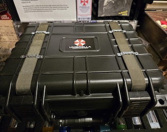 May include: A black, rectangular case with the Umbrella Corporation logo. The case has a textured surface, reinforced corners, and two olive green straps with metal clasps. The case is designed for secure storage and transport.