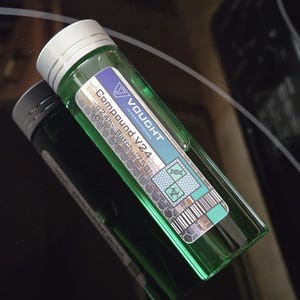 Può includere: Una fiala di vetro verde con tappo argentato, etichettata "Vought International Compound V24". L'etichetta include testo, un codice a barre e simboli di pericolo biologico. La fiala contiene un liquido verde e si riflette su una superficie nera.