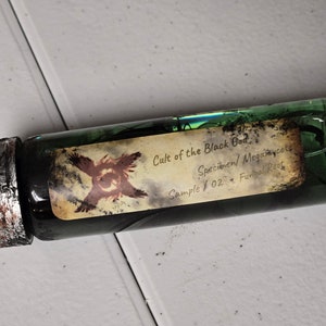 Puede incluir: Un vial de vidrio verde con una tapa de metal, que contiene un líquido oscuro y una etiqueta que dice "Cult of the Black God". La etiqueta también incluye el texto "Specimen/ Megamycete" y "Sample 02".
