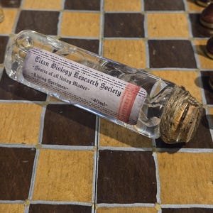 May include: A clear glass bottle with a cork stopper and a label that reads "Titan Biology Research Society - Source of all living Matter - 40mL - Living Specimen".