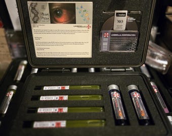 Puede incluir: Una caja negra con varios viales y un disco etiquetado como "UMBRELLA CORPORATION". Los viales están etiquetados como "G-VIRUS" y tienen un símbolo de riesgo biológico. El disco es un disco MO de 640 MB.