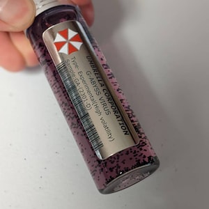 Op de afbeelding: Een klein, doorzichtig glazen flesje met een paarse vloeistof erin. Het flesje heeft een zwart etiket met de tekst "UMBRELLA CORPORATION G-ABYSS VIRUS Type- Experimental (High volatility) Sample: GA-(2301-D)" en een rood en wit biohazard-symbool.