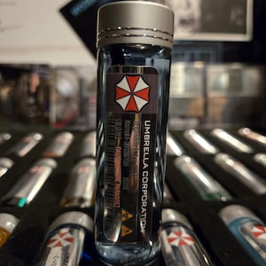 以下が含まれることがあります： 銀色のキャップが付いた透明なガラスバイアルで、「Umbrella Corporation」とラベル付けされています。 ラベルには赤と白の傘のロゴが付いています。 バイアルには青い液体が入っており、同様のバイアルのコレクションの一部です。