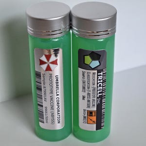 May include: Two small, clear plastic bottles with green liquid inside. The bottles have silver caps and labels with black text. The labels read "Umbrella Corporation Prototype Vaccine (Abyss)" and "Tricell Inc. Medication-[Prototype Vaccine] Classification-[T-Abyss Antigen] Sample-[TA7718922] 50ml Batch-[8832/C]".
