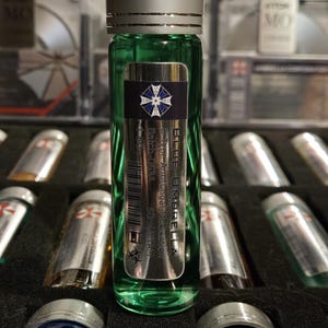 Puede incluir: Un vial de vidrio verde con una tapa plateada y una etiqueta metálica. La etiqueta dice "BLUE UMBRELLA" y presenta un logotipo azul y blanco. El vial está lleno de un líquido verde y forma parte de una colección de viales similares.