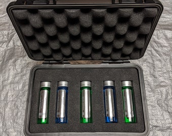 Pode incluir: Uma mala preta rígida aberta com acolchoamento de espuma contém seis frascos de vidro. Os frascos têm tampas e rótulos prateados, com líquido verde e azul no interior. A mala tem uma pega e duas fechaduras.