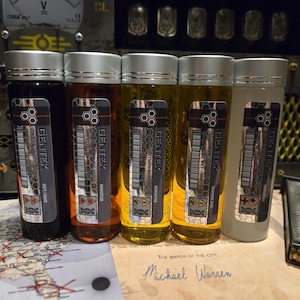 May include: Five glass bottles with silver lids, each containing a different colored liquid. The bottles have black and silver labels that read "GENTEK" and include a barcode. The liquids range from dark brown to clear. The text "HOPE/CAMO" is visible on one label.