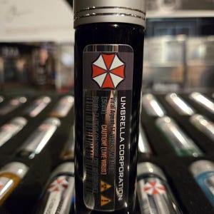 Op de afbeelding: Een zwarte flacon met een zilveren dop, met het opschrift "UMBRELLA CORPORATION". De flacon bevat een donkere vloeistof en een sticker met een rood en wit logo. De tekst op het etiket luidt "Viral Agent [T-VIRUS/MUTATED]" en "CAUTION! [LIVE VIRUS]" Andere flacons zijn zichtbaar.