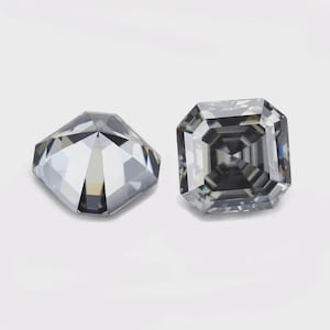 Op de afbeelding: Twee losse, gefacetteerde edelstenen met een vierkant, trapsgewijs geslepen ontwerp. De stenen hebben een doorschijnende, koelgrijze kleur met scherpe geometrische facetten die een contrastrijk spel van licht- en donkerreflecties creëren.