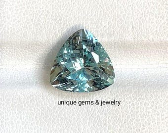 May include: A triangular, faceted gemstone with a light blue hue. The gem is set against a white background. The text "unique gems & jewelry" is below the gem.
