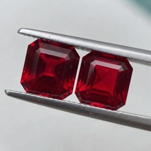 May include: Two square-cut, deep red gemstones held by silver-toned metal tweezers. The stones feature a faceted surface with a clear, transparent quality, reflecting light across their geometric edges and vibrant crimson color profile.