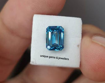 May include: A rectangular, faceted, blue gemstone. The gem is set on a white surface, with the text "unique gems & jewellers" visible. The gem has a vibrant, clear color and is likely a precious stone.