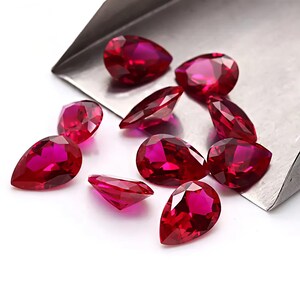 May include: A collection of faceted, teardrop-shaped rubies. The gems are a deep, vibrant red with multiple facets that catch the light. They are scattered on a white surface, with a silver tray in the background.