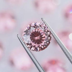 Op de afbeelding: Een ronde, gefacetteerde roze edelsteen wordt vastgehouden door een zilveren pincet. De edelsteen heeft een complexe interne structuur en is omgeven door andere onscherpe roze edelstenen. De achtergrond is zacht, wazig roze.