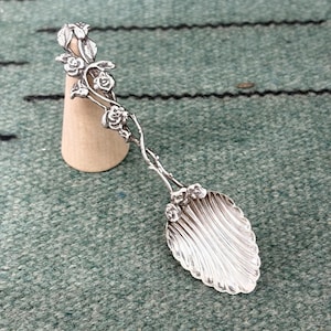 Puede incluir: Una cuchara de metal plateado con un mango decorado con rosas y hojas esculpidas. El cuenco de la cuchara presenta un diseño estriado en forma de concha con crestas verticales. La cuchara se apoya contra un pequeño soporte cónico claro.