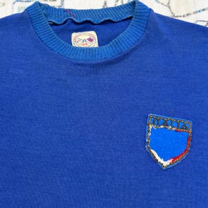 May include: A royal blue crewneck sweatshirt featuring a ribbed collar and a label at the neckline with the text GS. On the chest, there is a shield-shaped patch with gold beaded trim and the word ITALIA embroidered in black thread.