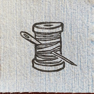 May include: A black and white illustration of a spool of thread and a needle on a blue denim background.