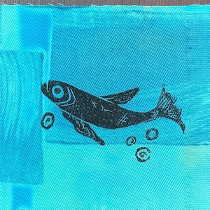 Puede incluir: Un parche de tela azul con una silueta negra de un pez nadando con burbujas a su alrededor.