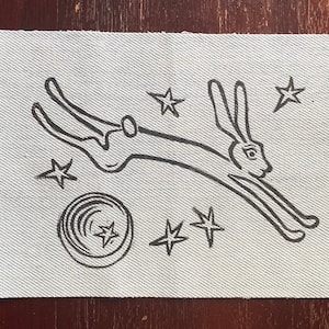 May include: A black and white illustration of a rabbit leaping in front of a crescent moon and stars. The rabbit is drawn in a simple, stylised manner.